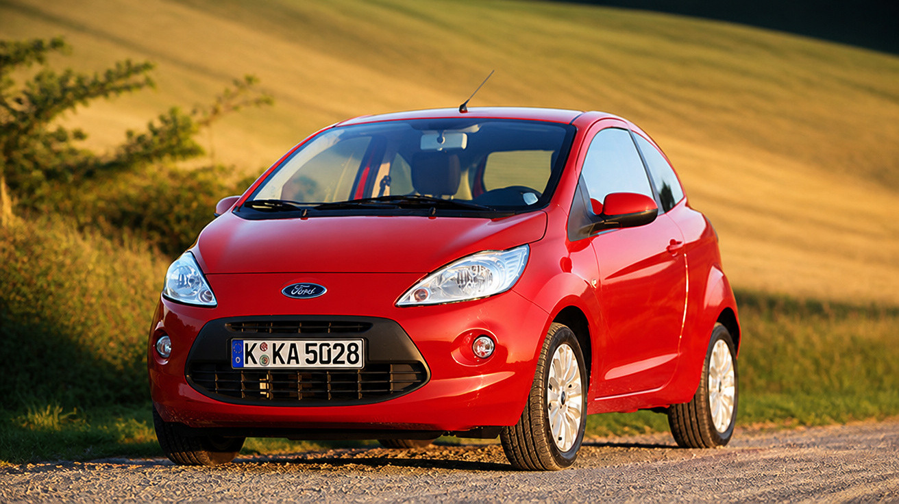 Roter Ford Kleinwagen steht auf einem Feldweg vor einem goldenen, hügeligen Hintergrund bei Sonnenlicht.