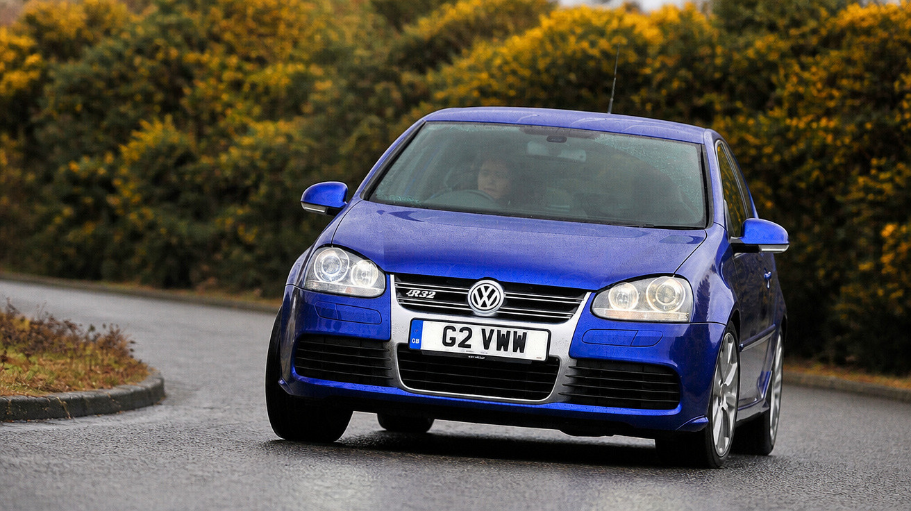 Blauer Volkswagen Golf fährt auf kurviger Straße, umgeben von grünen Büschen im Hintergrund.