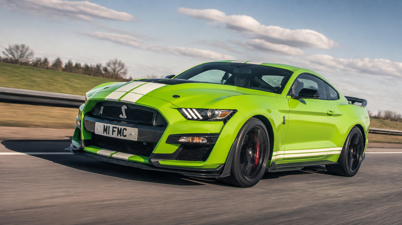Grüner Ford Mustang Shelby GT500 fährt auf einer Straße bei klarem Himmel mit weißen Rennstreifen.