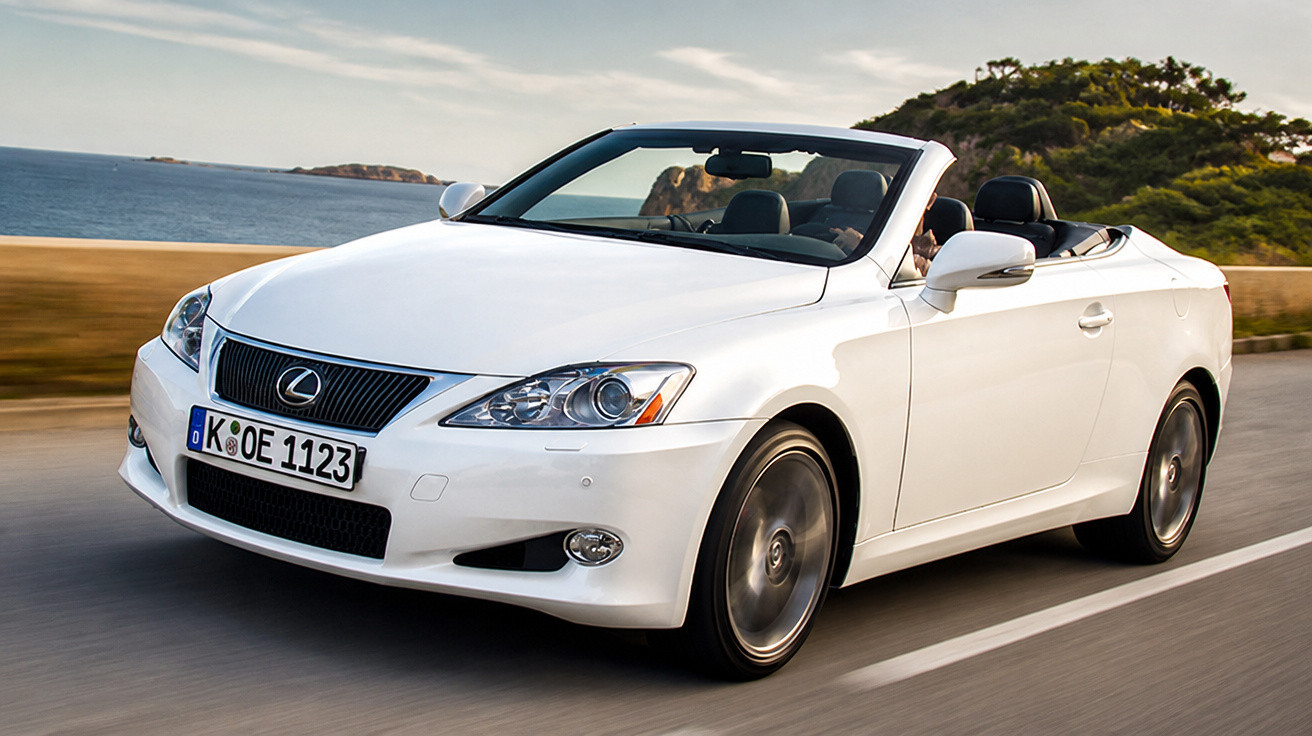 Weißer Lexus Cabrio fährt auf Küstenstraße bei sonnigem Wetter mit Meer und Hügeln im Hintergrund.