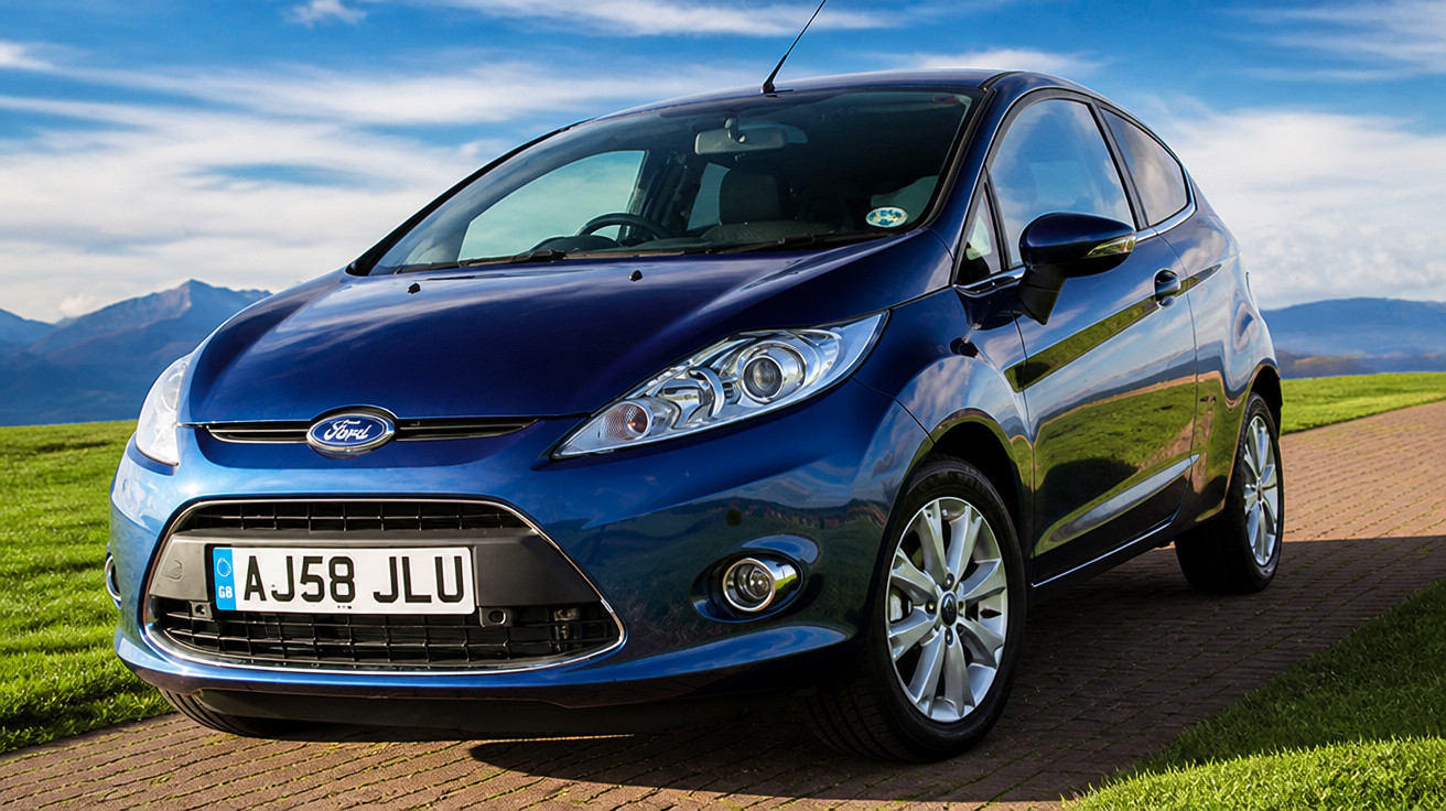 Blauer Ford Fiesta parkt auf Parkplatz mit Berglandschaft und blauem Himmel im Hintergrund.
