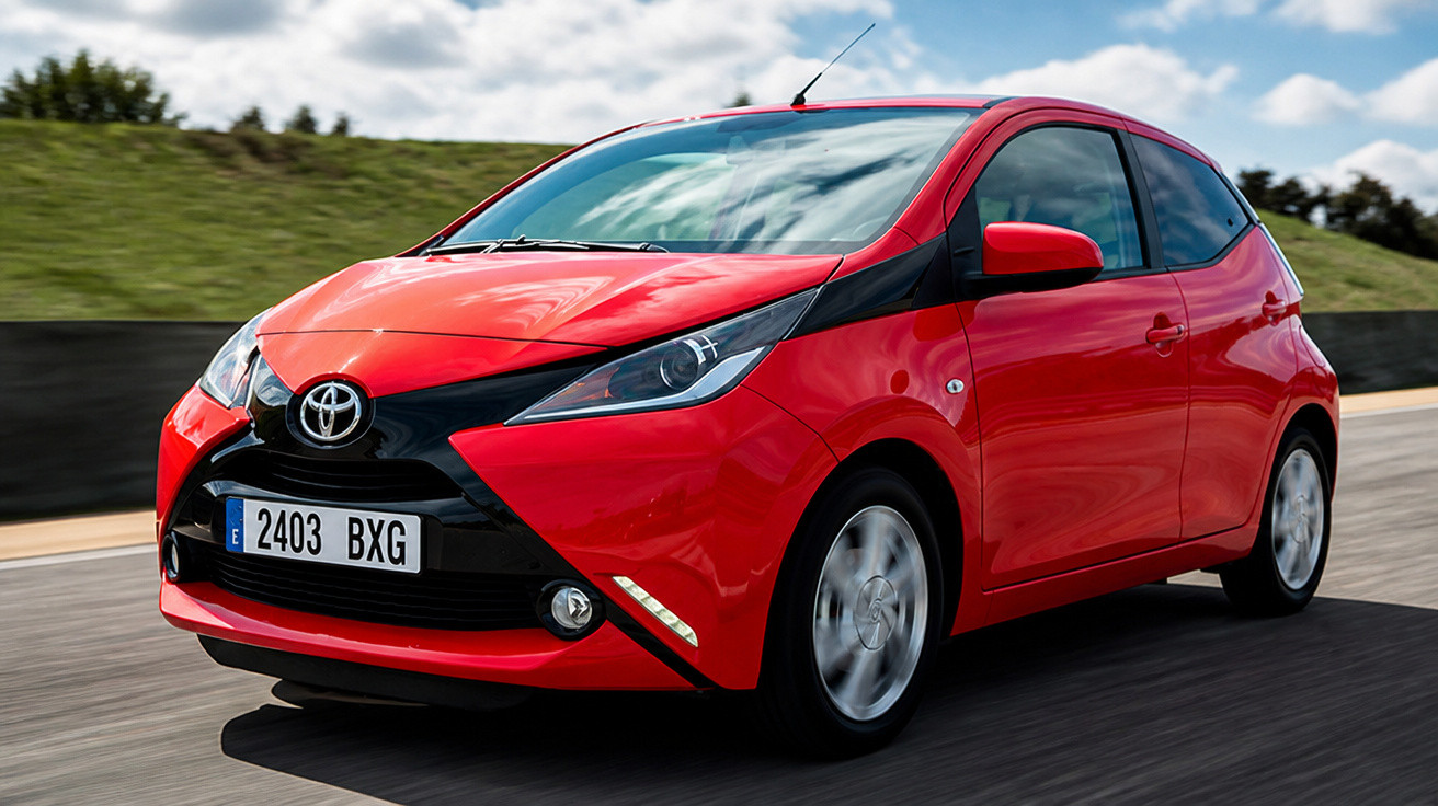 Roter Toyota Aygo fährt auf Asphaltstraße bei leicht bewölktem Himmel, Seitenansicht vorne rechts.