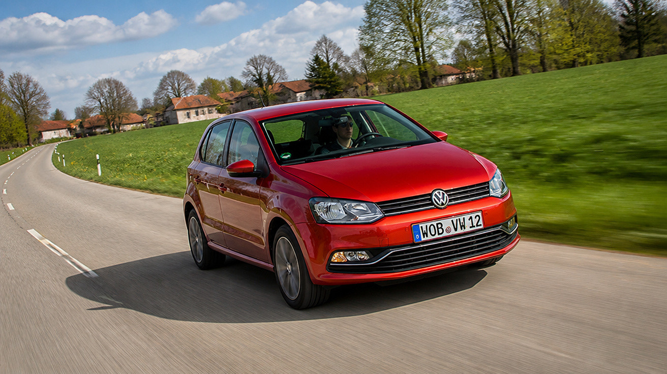 Roter Volkswagen Kleinwagen fährt auf Landstraße bei sonnigem Frühlingswetter mit Bäumen und Feldern.