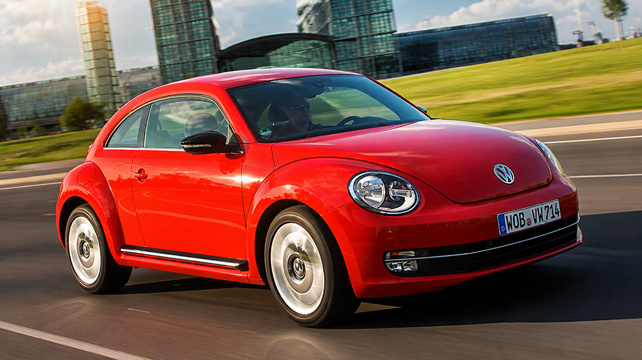 Roter Volkswagen Beetle fährt auf einer Straße vor modernen Glasgebäuden bei Tageslicht.