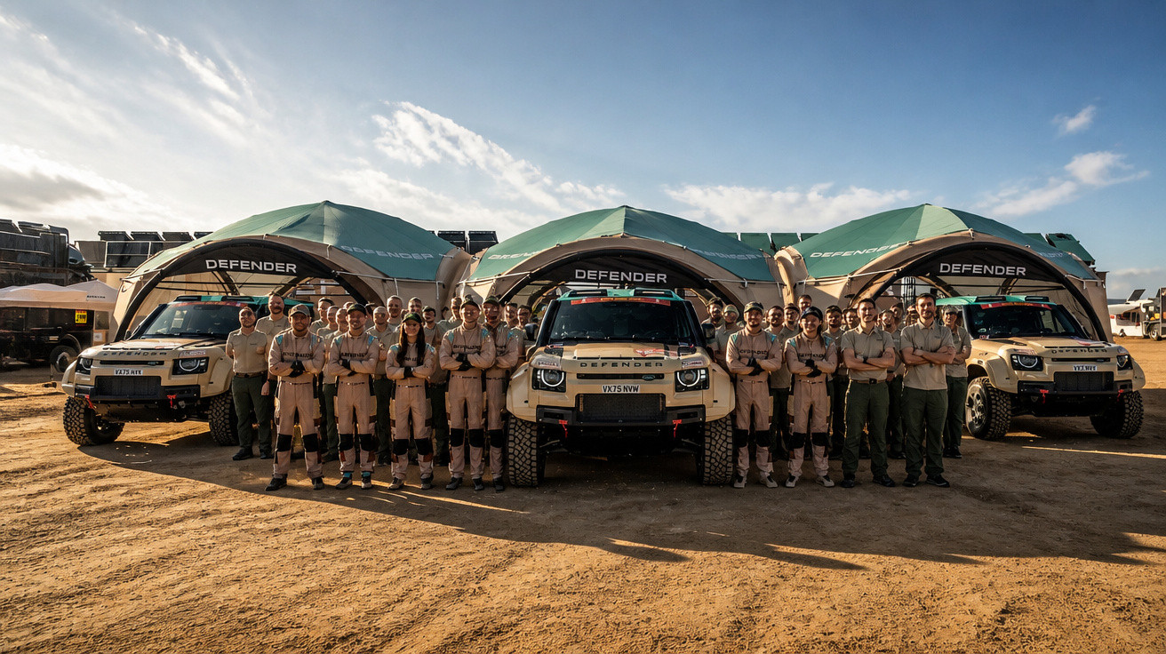 Gruppe von Personen in Arbeitskleidung vor drei Land Rover Defender Fahrzeugen und Zelten bei sonnigem Wetter.